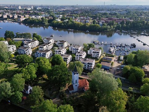 Aerial view of church spires piercing through lush green canopies, juxtaposed against modern apartments by the serene waters near Alt-Stralau, Berlin, Berlin, Germany.