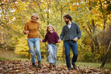 Fototapeta premium A cheerful family strolls through a park covered in autumn leaves. The parents and their child share laughter and joy while admiring the beautiful fall scenery around them.