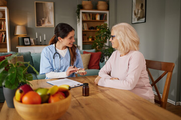 A home health nurse talks to a senior patient about medications while sitting at a table. The...