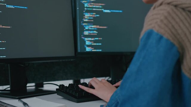 Close up of software engineer hands typing on mechanical keyboard with programming code on dual computer screens in dark office. Concept of AI development and digital innovation.