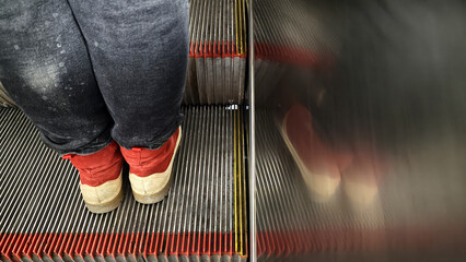 A person in worn pants and red shoes rides an escalator. The shot contrasts the sharp detail of the steps with a blurred reflection, creating an abstract, pensive mood. Urban, commuter concept © Анатолий Еремин