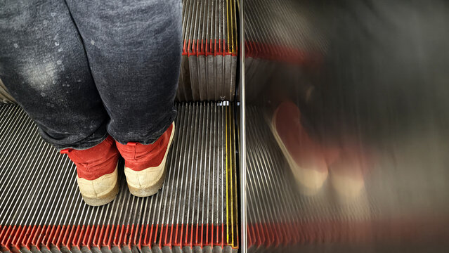A person in worn pants and red shoes rides an escalator. The shot contrasts the sharp detail of the steps with a blurred reflection, creating an abstract, pensive mood. Urban, commuter concept