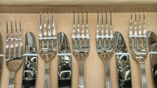 A new set of shiny silver cutlery, forks and knives, arranged neatly in a row inside a brown cardboard box. Top-down view with organized, repetitive patterns. Concept of dining, elegance, or a gift