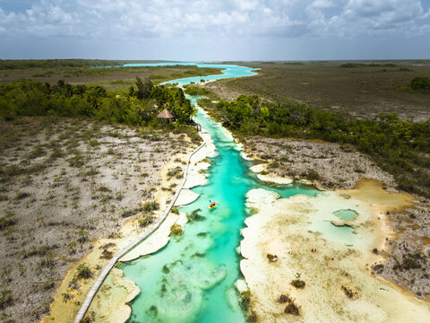 Aerial view of the vibrant turquoise river snakes through the landscape, contrasting with the surrounding lush greenery and sandy banks, Bacalar, Quintana Roo, Mexico.