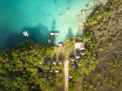 Aerial view of the vibrant turquoise waters meeting the shore with a dock, contrasted by the lush green jungle and rustic huts, Bacalar, Quintana Roo, Mexico.