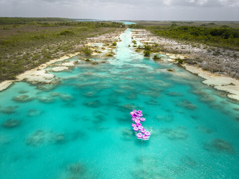 Aerial view of a turquoise lagoon with pink floaties nestled in the clear waters, contrasted by the vibrant green vegetation along the shore, Bacalar, Quintana Roo, Mexico.
