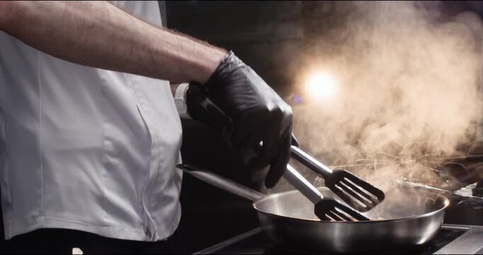 Master in gloves turns meat slices with tongs on frying pan in kitchen closeup. Skilled chef cooks beef dish in fancy restaurant. Professional food preparation