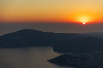 Fototapeta premium Tranquil sunset slides below dark distant mountains, casting warm evening light over a coastal valley and calm sea. Silhouettes and mist create a peaceful, scenic landscape. Turkey