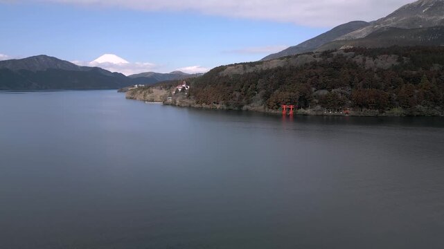 Scenic view of red torii shrine gate at Lake Ashi with snow-capped Mount Fuji in distance. Traditional Japanese landmark surrounded by forest mountains. Popular tourist spot in Hakone region.