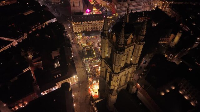 High-angle drone shot over the illuminated Ghent Belfry, capturing the bustling Christmas market below with crowds of people, glowing holiday stalls, and festive lights in Ghent, Belgium.
