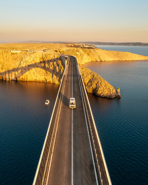Aerial view of a lone vehicle traverses the Pag Bridge, connecting the stark, sun-drenched Croatian island to the mainland, its golden cliffs meeting the deep blue Adriatic Sea, Pag, Croatia.