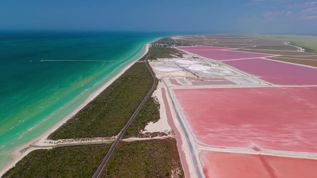 Aerial view of vibrant pink salt lakes contrasting with the turquoise sea along a sandy coastline, a road runs parallel to the shore, Las Coloradas, Yucatan, Mexico.