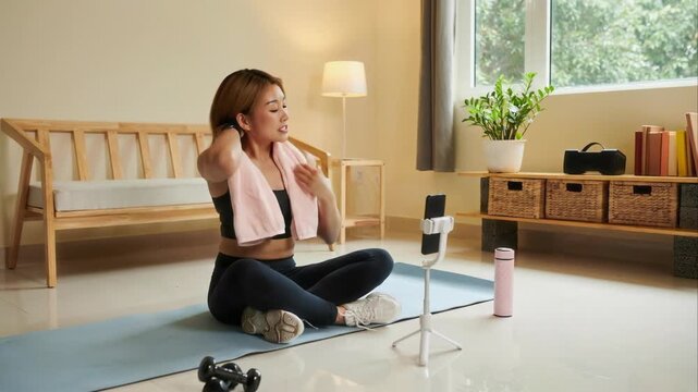 Full shot of confident young Asian female blogger in sportswear sitting on mat in modern living room and talking to smartphone camera on monopod while recording online training