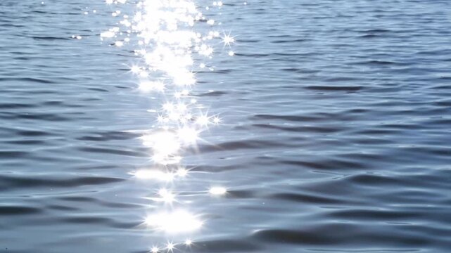 Sunlight sparkles on rippling lake water as bright reflections shimmer and drift with small waves, forming a moving glitter path across the blue surface in calm midday light, ideal background.