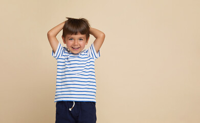 Frontal portrait of a happy little boy in a striped t-shirt, posing in the studio, making faces, having fun, on a beige background. Copy space.