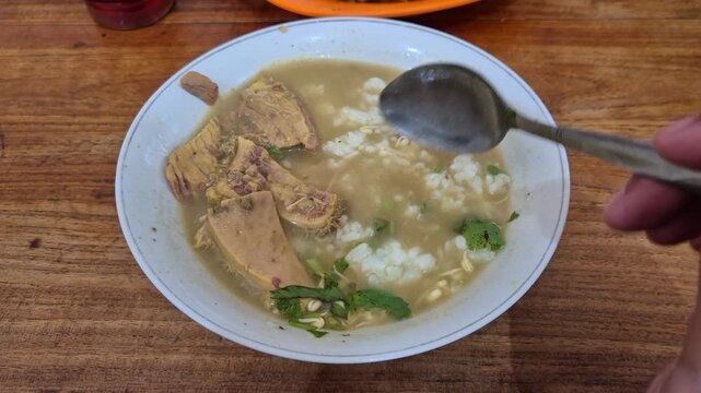 a bowl of traditional indonesian soup called soto daging containing sliced block of meat, rice, some spices, and broth  mixed with coconut milk while stirred using metal spoon.