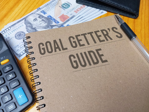 Brown spiral notebook with GOAL GETTER'S GUIDE text on a wooden desk with a calculator and dollar bills. Success planning concept.