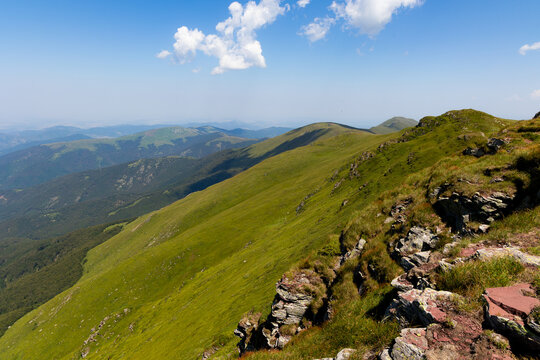 View of rolling green hills meet rocky terrain under a vast blue sky dotted with clouds, creating a serene yet rugged landscape, Bela Palanka, Pirotski Okrug, Serbia.