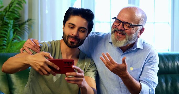 Indian young son and senior father taking selfie together using smartphone while sitting on sofa in modern home, capturing happy family moment and freezing joyful memory between two generations