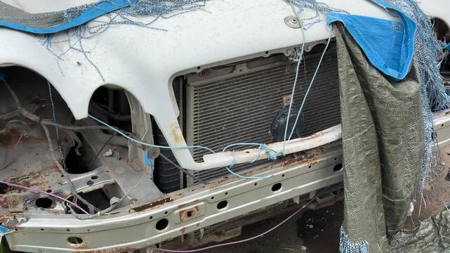 Dismantled wrecked sedan front end exposed, blue tarp draped over mangled grille, missing headlights, twisted bumper and exposed radiator, weeds and grass encroaching on abandoned vehicle.
