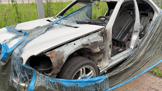 Dismantled wrecked sedan front end exposed, blue tarp draped over mangled grille, missing headlights, twisted bumper and exposed radiator, weeds and grass encroaching on abandoned vehicle.