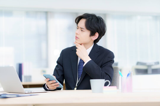 オフィスでノートパソコンで仕事をする困った顔のスーツを着た若い日本人男性