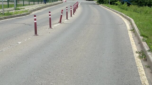Curved asphalt road lined with bollards, red white plastic posts separating lane from shoulder, faded yellow edge paint and concrete curb, quiet suburban stretch with green verge, durable.
