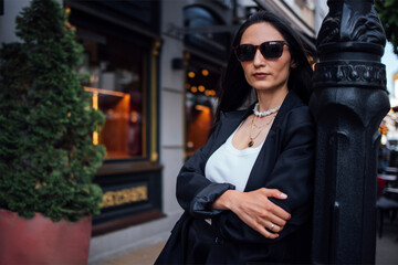 Obraz premium Stylish woman with long dark hair wearing sunglasses and a black blazer, leaning against a lamppost, exuding confidence and elegance in an urban setting with greenery