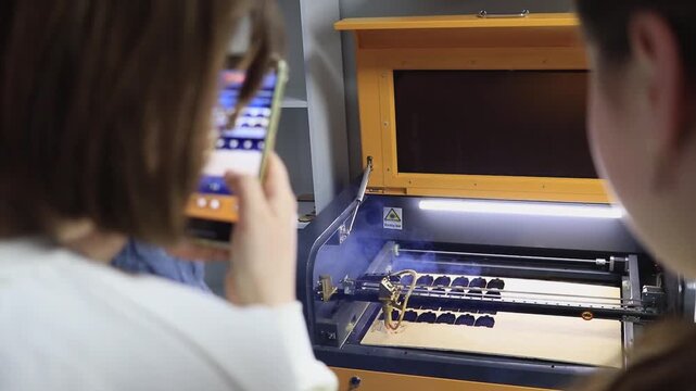 University students undergo a practical training in the digital manufacturing laboratory. Operation of the laser machine and recording the process on a smartphone during technical training.