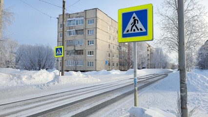 Pedestrian crossing sign on a snowy city street against the backdrop of a typical residential panel building in winter. Road covered with snow and ice rime on trees under a clear blue sky. Russian urb © Олег Шерстнев