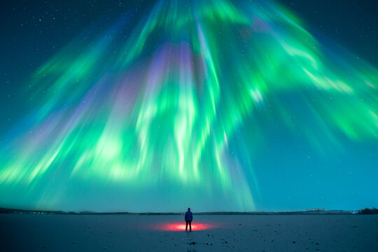 Night winter scene with lone man using red flashlight on snow-covered field beneath aurora borealis and star-filled sky, polar lights, northern lights, northern wilderness