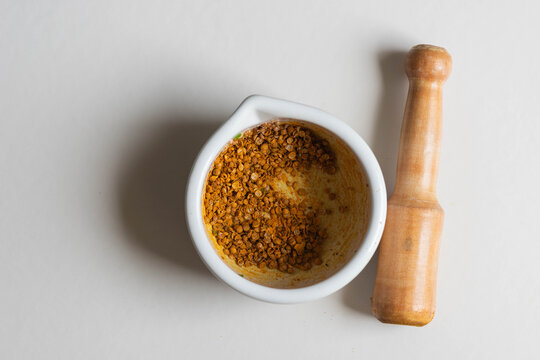 Mortero blanco de cer&aacute;mica con semillas de cilantro machacadas o majadas. A su lado mazo de madera. Plano cenital. 