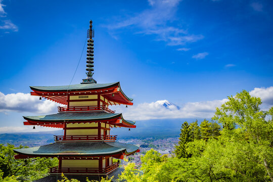 View of a red pagoda against a vibrant blue sky, with Mount Fuji a ghostly presence in the background, surrounded by lush green trees, Fujiyoshida, Yamanashi, Japan.
