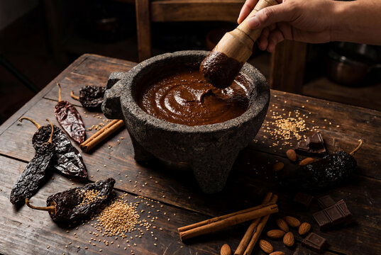 Hand using wooden pestle to mix traditional Mexican mole sauce in stone molcajete mortar with chili pepper cinnamon and chocolate on rustic table