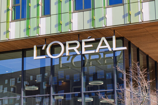 Close-up of the white L'Oreal logo on the glass facade of the corporate headquarters in Hoofddorp, Netherlands, under a clear blue sky.