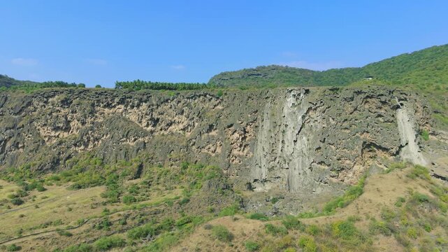 Aerial view of a massive limestone cliff face with a row of palm trees on the plateau and green shrubs below in Salalah, Oman.