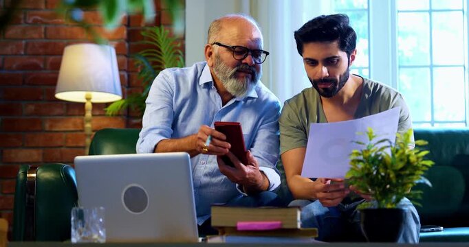 Indian retired father and young son working together on laptop and smartphone at home, reviewing financial report, share trading data and online business documents while discussing profit and loss