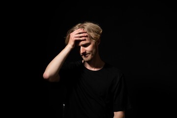 Young man experiencing headache or regret, holding hand to forehead, feeling pain, stress, or despair, standing on a dark background