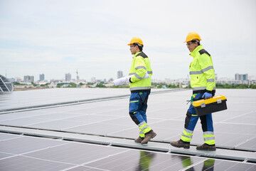 Two construction workers walking on a rooftop solar panel array, one carrying a blueprint, another with a toolbox. They wear safety gear, indicating a professional environment