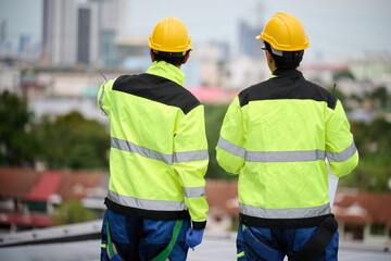 Back view engineers on a rooftop, inspecting a solar panel installation, with a cityscape in the background. They wear safety vests and hard hats while reviewing a blueprint.