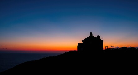 Obraz premium Bright summer sky blending blue and orange colors behind a flawless silhouette of a charming building with sea scenery background