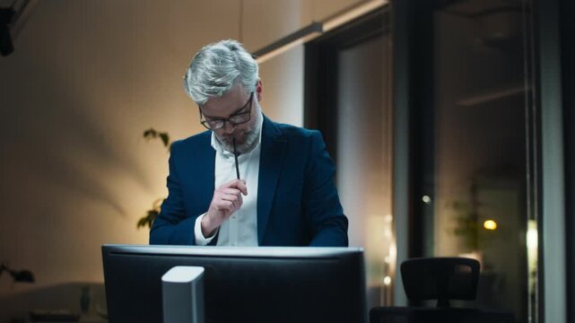 Caucasian man standing at workstation. Mature male manager holding clipboard and pencil. Business professional thinking about solution. Computer monitor in front. Nighttime office setting.