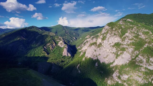 Aerial pan revealing sunlit rocky Altai mountain ridge and deep forested valley with shifting shadows, flying over alpine peaks for travel and nature background.