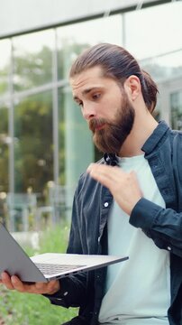 Successful bearded businessman greeting colleagues with hand gesture during online conference on laptop outdoors, working remotely. Concept of modern flexible work and global digital communication