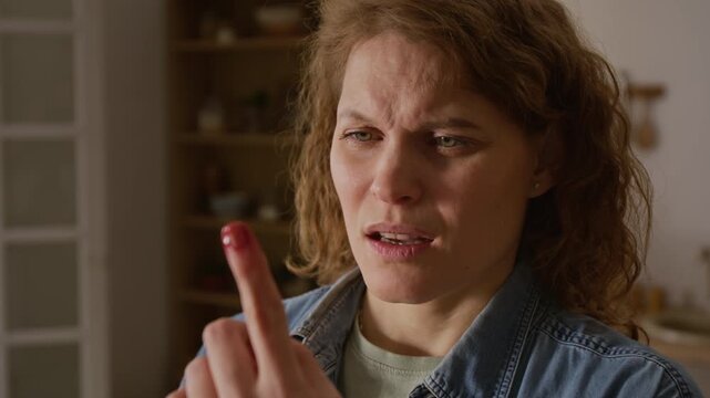 Close-up of face of shocked mature Caucasian woman cutting herself while chopping vegetables for cooking, then staring at bloody finger, putting it in mouth to stem bleeding