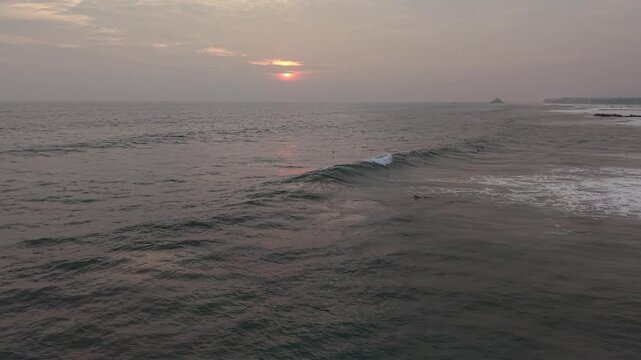 Surfers riding ocean waves during a golden sunset at Naomi Beach in Sri Lanka. Silhouettes of surfers against dramatic orange sky, tropical coastline, peaceful evening atmosphere, and authentic island