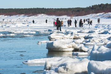 Icy winter seascape with frozen waves, floating ice floes on the blue sea water along a snowy shoreline and people walking on the Baltic Sea in Palanga, Lithuania