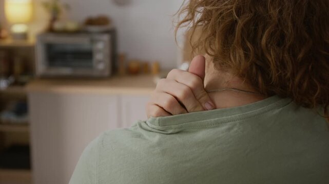 Rear close-up of hands of anonymous female rubbing stiff shoulder and neck, to relieve muscle tension, while suffering from chronic pain while doing mundane daily activities