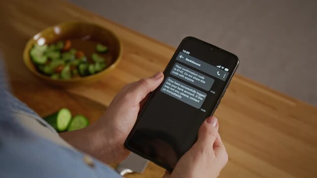 Over-shoulder close-up of female hands holding smartphone with bank messages displayed on screen, password and login notifications, while cooking in kitchen, suggesting cybercrime and fraud