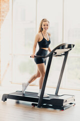 Fototapeta premium Young woman exercises on a treadmill in a bright gym with modern equipment and comfortable surroundings, enjoying her fitness routine